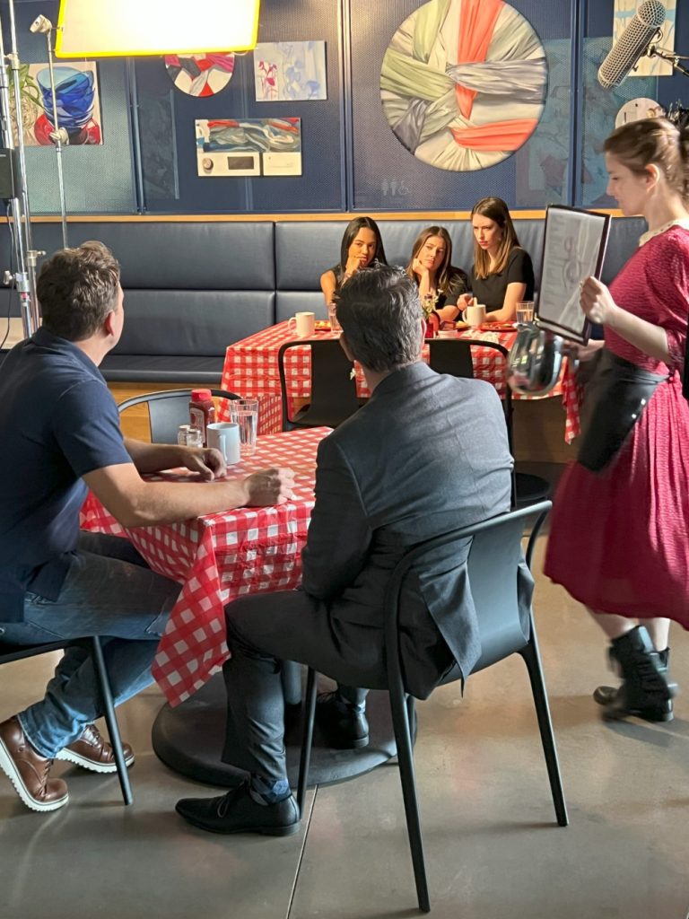 Two male actors in the foreground, with one female actor standing; three actresses in the background. All sitting around tables with red checked tablecloths.